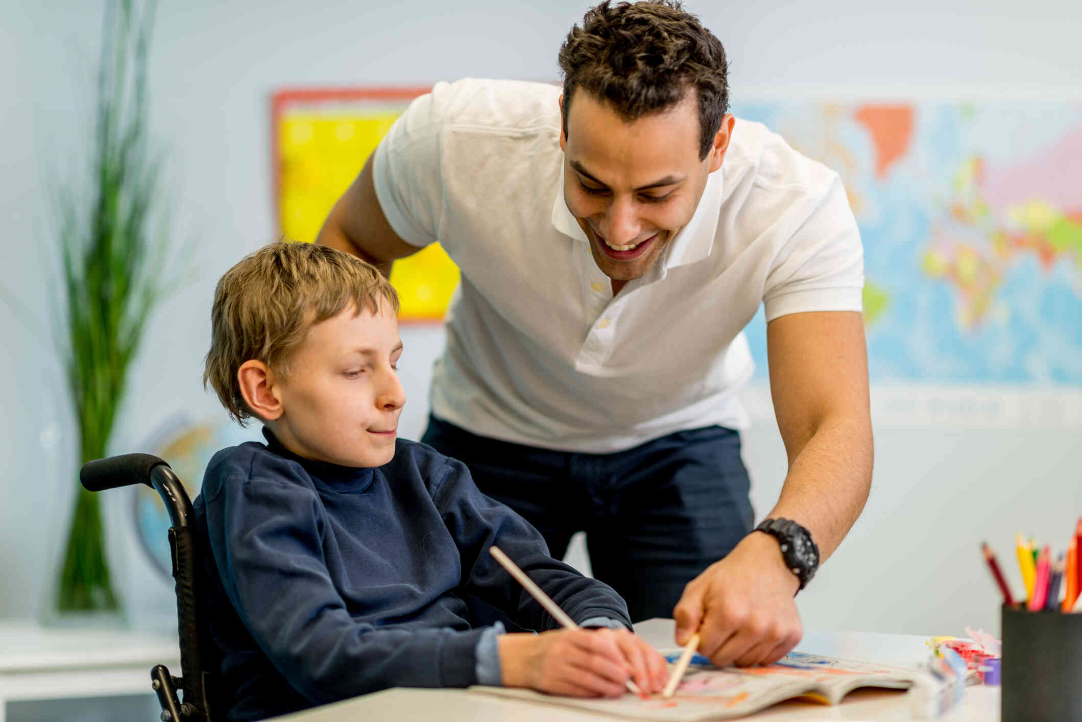 A young boy in a wheelchair sits at a table writting in a book as a man leans over to help him while smiling.